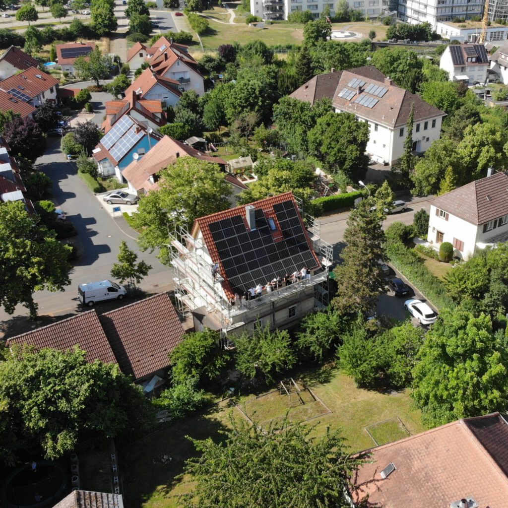 A house under renovation is surrounded by scaffolding, with solar panels installed on its roof; trees and other buildings are visible in the background.